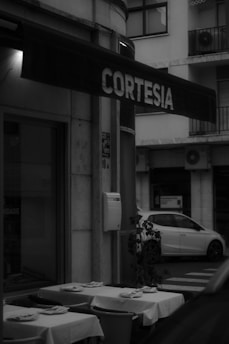 A black and white photo of a small outdoor restaurant area with neatly arranged tables and chairs. Each table is covered with a tablecloth and set with napkins. There is an overhanging sign with the word 'Cortesía' written on it. A potted plant is adjacent to a white car parked on the street, with an apartment building in the background.