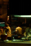 A dimly lit kitchen scene where three chefs are actively preparing food. There is a warm, ambient glow from the overhead lights. One chef is cooking at the stove, while another is working with utensils beside him. The kitchen is equipped with various utensils and ingredients, and there is a visible monitor displaying data or orders.