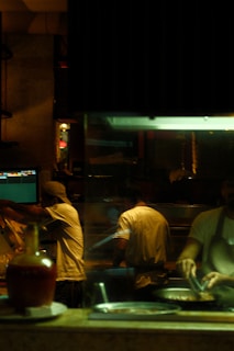A dimly lit kitchen scene where three chefs are actively preparing food. There is a warm, ambient glow from the overhead lights. One chef is cooking at the stove, while another is working with utensils beside him. The kitchen is equipped with various utensils and ingredients, and there is a visible monitor displaying data or orders.