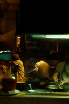 A dimly lit kitchen scene where three chefs are actively preparing food. There is a warm, ambient glow from the overhead lights. One chef is cooking at the stove, while another is working with utensils beside him. The kitchen is equipped with various utensils and ingredients, and there is a visible monitor displaying data or orders.