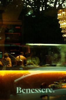 The image captures the interior of a restaurant or cafe, with the word 'Benessere' prominently displayed on a reflective surface. There are people working behind a counter, possibly chefs or kitchen staff, with a warm light illuminating the area. A chandelier with multiple hanging lights adds elegance, and there are some tables set for dining in the foreground.