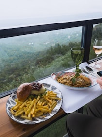 A meal is arranged on a table with a scenic, green landscape visible through a window. The setting includes a plate with a cheeseburger topped with caramelized onions, accompanied by French fries. Another plate holds pasta garnished with grated cheese and basil. Two glasses are present, one with a green tint and another containing a pinkish beverage, likely wine. The atmosphere is relaxed and inviting, offering a combination of good food and a picturesque view.