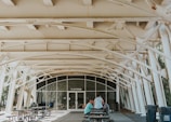 An outdoor covered space with white metal beams and a restroom entrance in the background. There are several picnic tables and benches, with a few people sitting and standing near them. The area is well-lit with natural light and there is greenery visible through the structural openings.