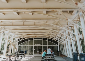 An outdoor covered space with white metal beams and a restroom entrance in the background. There are several picnic tables and benches, with a few people sitting and standing near them. The area is well-lit with natural light and there is greenery visible through the structural openings.