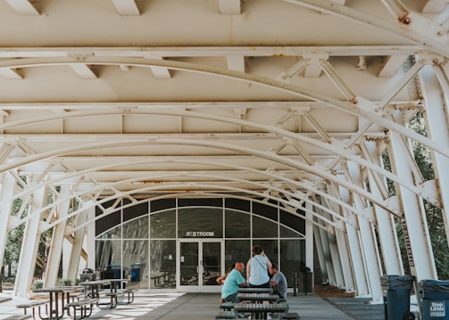 An outdoor covered space with white metal beams and a restroom entrance in the background. There are several picnic tables and benches, with a few people sitting and standing near them. The area is well-lit with natural light and there is greenery visible through the structural openings.