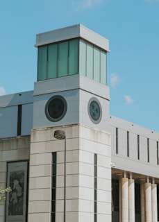A modern building with a prominent tower section featuring large windows and circular emblems. The structure is made of light-colored stone with tall columns on one side and reflects a contemporary architectural style.