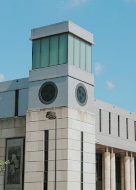 A modern building with a prominent tower section featuring large windows and circular emblems. The structure is made of light-colored stone with tall columns on one side and reflects a contemporary architectural style.