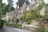 A row of charming stone cottages with steeply pitched roofs and chimneys, adorned with climbing plants and surrounded by lush greenery. The cottages are situated along a narrow, winding road lined with a low stone wall and various shrubs and trees.