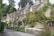 A row of charming stone cottages with steeply pitched roofs and chimneys, adorned with climbing plants and surrounded by lush greenery. The cottages are situated along a narrow, winding road lined with a low stone wall and various shrubs and trees.
