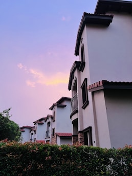 A row of modern residential houses with red-tiled roofs and arched windows is seen against a backdrop of a colorful, pastel evening sky. The foreground features lush green hedges, adding a touch of nature to the scene.