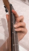 A hand is positioned on the fretboard of a guitar, fingers pressing down on the strings to form a chord. The neck of the guitar is made of wood, and the strings are metallic, offering a close-up view of the instrument and hand interaction.