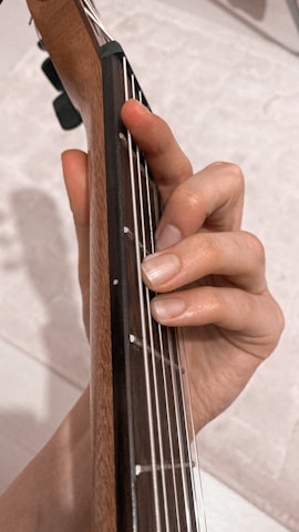A hand is positioned on the fretboard of a guitar, fingers pressing down on the strings to form a chord. The neck of the guitar is made of wood, and the strings are metallic, offering a close-up view of the instrument and hand interaction.