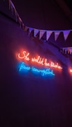 Neon lights illuminate a dark wall with a cursive message in red and blue. Above the lights, a string of triangular flags or bunting hangs, adding a festive element.