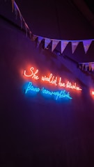 Neon lights illuminate a dark wall with a cursive message in red and blue. Above the lights, a string of triangular flags or bunting hangs, adding a festive element.