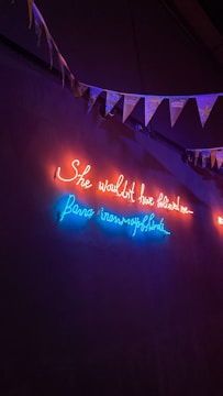 Neon lights illuminate a dark wall with a cursive message in red and blue. Above the lights, a string of triangular flags or bunting hangs, adding a festive element.