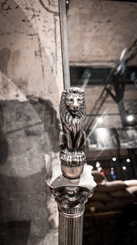 A metallic sculpture of a lion stands elegantly on a narrow column. The background appears to be a dimly lit, rustic interior with a mixture of industrial and antique elements. The surface on which the lion rests is ornate and intricately designed.