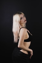 A person with platinum blonde hair poses against a dark background, wearing a black top and dark pants. The lighting creates a dramatic contrast, highlighting their features and the smooth texture of their hair. Their expression is composed and confident, with a slight tilt of the head enhancing the pose.