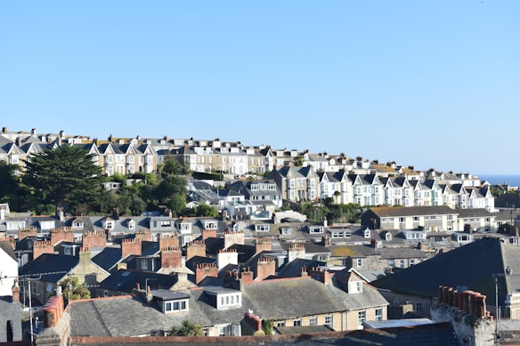 A scenic view of a seaside town, featuring rows of traditional terraced houses. The homes are tightly packed and adorned with chimneys, situated on a hill that slopes gently toward the sea. The sky is clear and blue, highlighting the peaceful and sunny atmosphere of the scene.