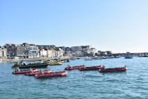 A coastal townscape with small red motorboats floating in a calm blue harbor. Behind the boats, there's a row of charming, traditional buildings with a mix of stone and white facades. The scene is set under a clear blue sky, creating a serene atmosphere.