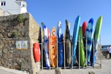 A row of colorful kayaks and surfboards is arranged vertically against a stone wall. The stone wall has some plants growing on it, and two signs indicating 'Danger' are visible. A red life ring is attached to the wall. Above the wall, part of a building can be seen with windows. The sky is clear and blue, hinting at a sunny day.