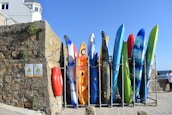 A row of colorful kayaks and surfboards is arranged vertically against a stone wall. The stone wall has some plants growing on it, and two signs indicating 'Danger' are visible. A red life ring is attached to the wall. Above the wall, part of a building can be seen with windows. The sky is clear and blue, hinting at a sunny day.