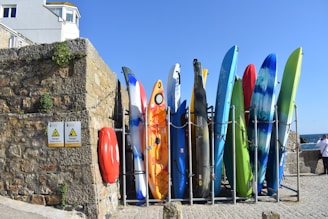 A row of colorful kayaks and surfboards is arranged vertically against a stone wall. The stone wall has some plants growing on it, and two signs indicating 'Danger' are visible. A red life ring is attached to the wall. Above the wall, part of a building can be seen with windows. The sky is clear and blue, hinting at a sunny day.