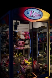A claw machine filled with various plush toys. The machine is illuminated with neon lights, giving it a vibrant and playful appearance. The sign at the top reads 'E Claw' with bright colors, and the glass showcases a variety of stuffed animals, including unicorns, bears, and other assorted characters and toys.