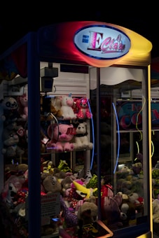 A claw machine filled with various plush toys. The machine is illuminated with neon lights, giving it a vibrant and playful appearance. The sign at the top reads 'E Claw' with bright colors, and the glass showcases a variety of stuffed animals, including unicorns, bears, and other assorted characters and toys.