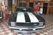 A vintage black car with white racing stripes is parked indoors. It features a 'Z28' emblem on the grille and is surrounded by retro decor, including a Gulf gas pump and a Coca-Cola vending machine. The floor has a checkered tile pattern and the walls display various memorabilia.
