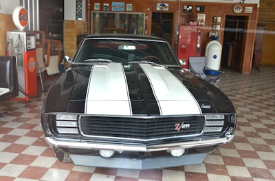 A vintage black car with white racing stripes is parked indoors. It features a 'Z28' emblem on the grille and is surrounded by retro decor, including a Gulf gas pump and a Coca-Cola vending machine. The floor has a checkered tile pattern and the walls display various memorabilia.