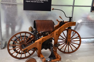 A vintage motorcycle with a wooden frame and large spoked wheels is displayed in a glass-enclosed space. The seat and various parts of the motorcycle are leather-padded. There are informational plaques on the walls and some reflections visible on the glass.