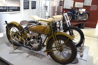 A classic Harley-Davidson motorcycle is displayed in a museum setting. The bike features an olive green and cream color scheme with visible mechanical components, including a large engine and vintage headlamps. Adjacent, another motorcycle with a blue color scheme is partially visible. The exhibit includes informational posters and blueprints on the walls.