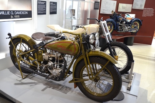 A classic Harley-Davidson motorcycle is displayed in a museum setting. The bike features an olive green and cream color scheme with visible mechanical components, including a large engine and vintage headlamps. Adjacent, another motorcycle with a blue color scheme is partially visible. The exhibit includes informational posters and blueprints on the walls.