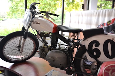 A vintage motorcycle is displayed indoors, featuring a white and red gas tank with the Harley Davidson logo. The number 60 is prominently shown on a race plate. The motorcycle's design includes an exposed engine, a leather seat with visible springs, and classic spoked wheels. Natural light enters through large windows in the background, highlighting the motorcycle's details.
