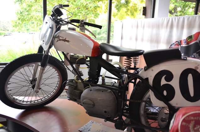 A vintage motorcycle is displayed indoors, featuring a white and red gas tank with the Harley Davidson logo. The number 60 is prominently shown on a race plate. The motorcycle's design includes an exposed engine, a leather seat with visible springs, and classic spoked wheels. Natural light enters through large windows in the background, highlighting the motorcycle's details.