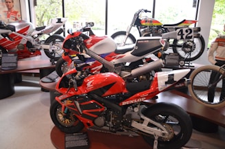 Several racing motorcycles are displayed indoors on platforms. The nearest bike is primarily red with black and white accents, bearing the brand Honda and featuring sponsor decals. It is surrounded by other bikes, including one with the number 42, showcasing a variety of designs and colors. A figure in a motorcycle jacket is visible to the right, standing beside the exhibits. The background includes large windows revealing a view of greenery outside.