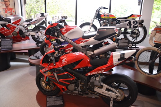 Several racing motorcycles are displayed indoors on platforms. The nearest bike is primarily red with black and white accents, bearing the brand Honda and featuring sponsor decals. It is surrounded by other bikes, including one with the number 42, showcasing a variety of designs and colors. A figure in a motorcycle jacket is visible to the right, standing beside the exhibits. The background includes large windows revealing a view of greenery outside.