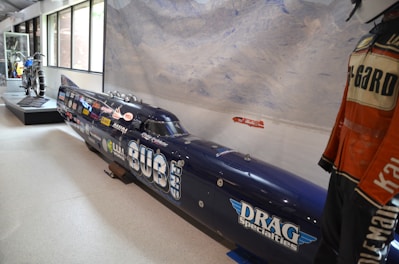 A display of a dark-colored land speed racing car adorned with multiple sponsor logos. The car is elongated and streamlined, positioned in an indoor setting with large windows on one side. To the left of the car is a motorcycle on display and to the right, a detailed view of a racing suit on a mannequin. The background features an artistic depiction of a mountainous landscape.