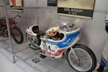 A vintage motorcycle with racing decals and a streamlined body is displayed inside a metal mesh enclosure. The motorcycle features a prominent number '104' and is branded with 'Harley Hospital' and 'Leo Payne-Pilot'. It has a small windshield and racing-style seat. Nearby, another motorcycle is partially visible. A plaque indicating 'Earl Bowlby' is mounted above.
