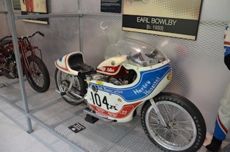A vintage motorcycle with racing decals and a streamlined body is displayed inside a metal mesh enclosure. The motorcycle features a prominent number '104' and is branded with 'Harley Hospital' and 'Leo Payne-Pilot'. It has a small windshield and racing-style seat. Nearby, another motorcycle is partially visible. A plaque indicating 'Earl Bowlby' is mounted above.