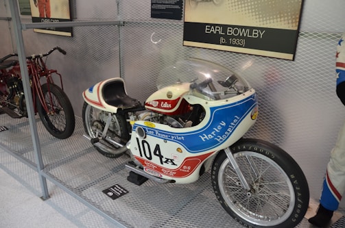 A vintage motorcycle with racing decals and a streamlined body is displayed inside a metal mesh enclosure. The motorcycle features a prominent number '104' and is branded with 'Harley Hospital' and 'Leo Payne-Pilot'. It has a small windshield and racing-style seat. Nearby, another motorcycle is partially visible. A plaque indicating 'Earl Bowlby' is mounted above.