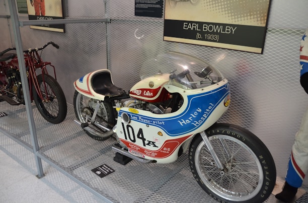 A vintage motorcycle with racing decals and a streamlined body is displayed inside a metal mesh enclosure. The motorcycle features a prominent number '104' and is branded with 'Harley Hospital' and 'Leo Payne-Pilot'. It has a small windshield and racing-style seat. Nearby, another motorcycle is partially visible. A plaque indicating 'Earl Bowlby' is mounted above.