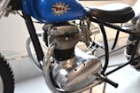 Close-up view of a classic motorcycle engine featuring a prominent chrome finish. The engine has a distinctive vintage design with several metallic components visible, including a prominent exhaust pipe. The blue fuel tank is labeled with a logo and the motorcycle frame is black. There is a thick treaded tire partially visible.