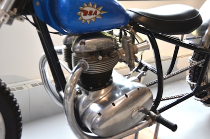 Close-up view of a classic motorcycle engine featuring a prominent chrome finish. The engine has a distinctive vintage design with several metallic components visible, including a prominent exhaust pipe. The blue fuel tank is labeled with a logo and the motorcycle frame is black. There is a thick treaded tire partially visible.