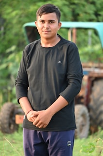 A young person stands outdoors with greenery in the background. They are wearing a black long-sleeved shirt and blue pants, and they have short dark hair. A partially visible tractor is in the background.