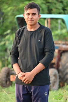 A young person stands outdoors with greenery in the background. They are wearing a black long-sleeved shirt and blue pants, and they have short dark hair. A partially visible tractor is in the background.