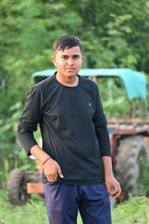 A young person stands outdoors, wearing a black long-sleeve shirt and dark pants. Their expression is neutral, and they are positioned slightly to the side with greenery and a hint of an old tractor in the background.