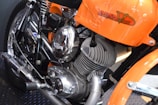 A close-up view of a motorcycle engine, prominently displaying a polished metallic finish. The frame and elements around the engine are colored in bright orange. The motorcycle brand name is visible on the orange part of the frame. The engine's design includes visible coils and exhaust pipes.