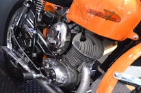 A close-up view of a motorcycle engine, prominently displaying a polished metallic finish. The frame and elements around the engine are colored in bright orange. The motorcycle brand name is visible on the orange part of the frame. The engine's design includes visible coils and exhaust pipes.