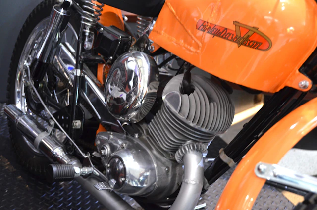 A close-up view of a motorcycle engine, prominently displaying a polished metallic finish. The frame and elements around the engine are colored in bright orange. The motorcycle brand name is visible on the orange part of the frame. The engine's design includes visible coils and exhaust pipes.